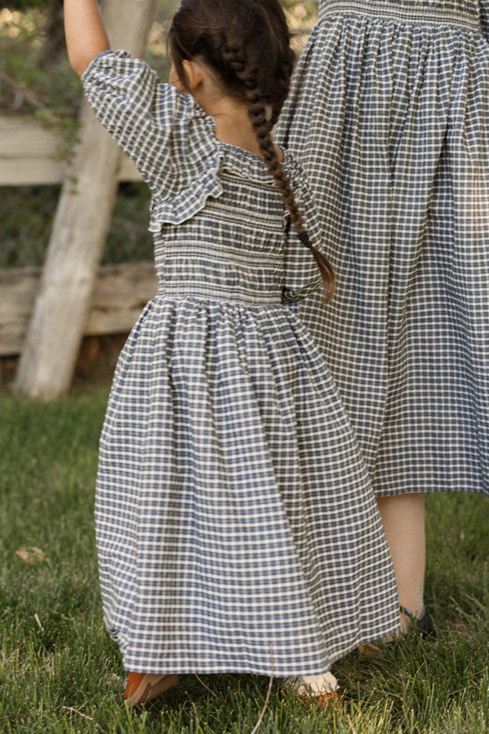 A young girl with a braid, dressed in the Ivy City Co Mini Mae 3/4 Sleeve Dress in Prussian Blue Plaid featuring a smocked bodice, stands on grass beside another person wearing a matching dress. Both are outdoors near a wooden fence.