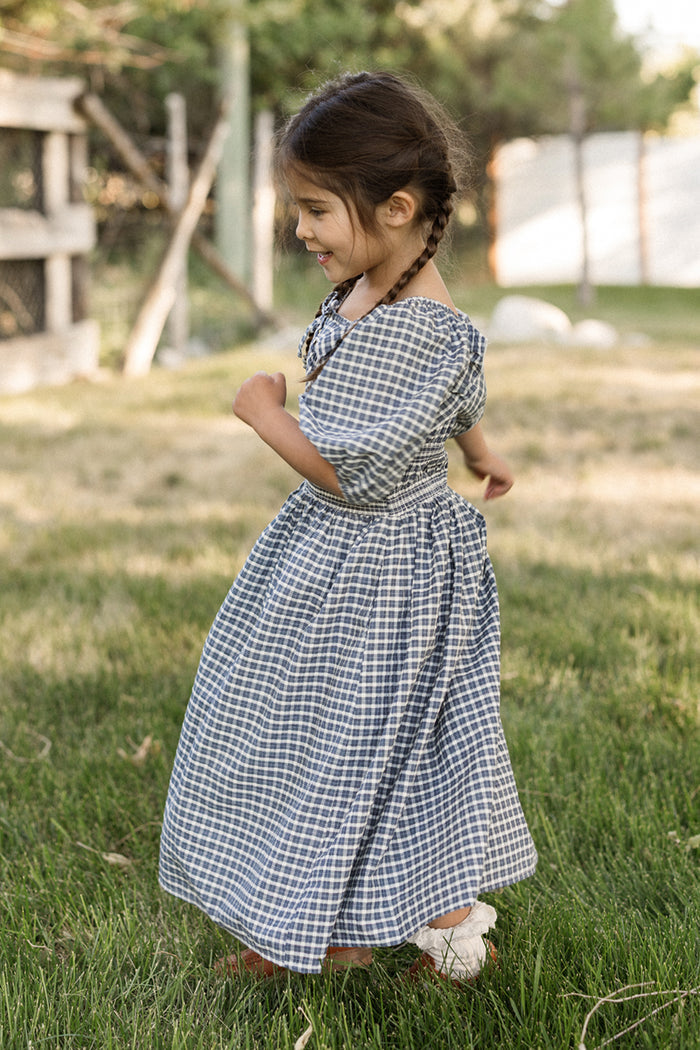 A young girl with braids spins playfully on green grass outdoors, smiling and enjoying a sunny day while wearing the Ivy City Co Mini Mae 3/4 Sleeve Dress in Prussian Blue Plaid, featuring a smocked bodice.