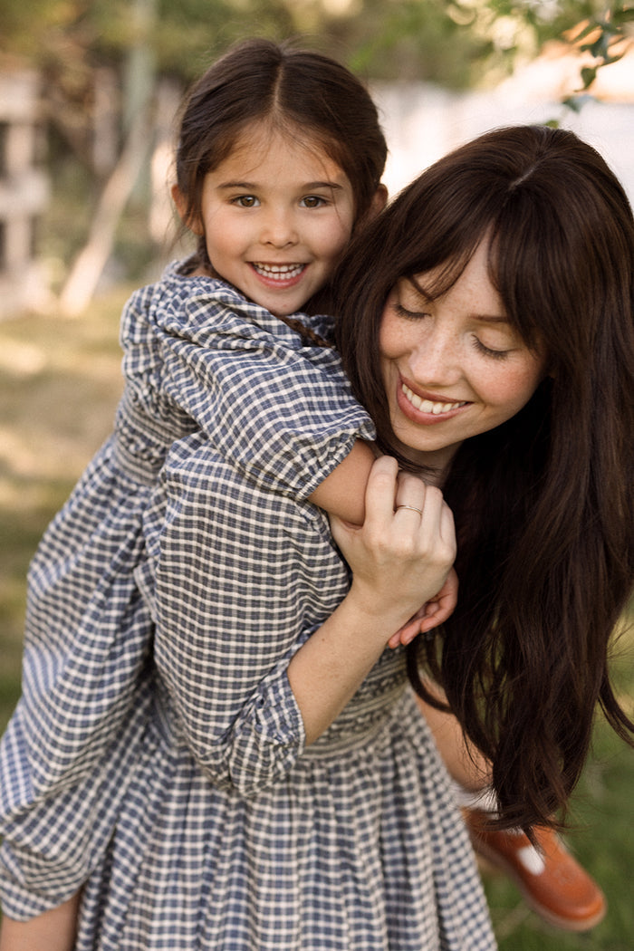 A woman with long brown hair gives a piggyback ride to a young, smiling girl. Both wear matching Ivy City Co Mini Mae 3/4 Sleeve Dresses in Prussian Blue Plaid, featuring charming smocked bodices, as they enjoy their time outdoors in a sunlit, grassy area.