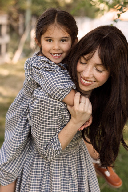 A woman with long brown hair gives a piggyback ride to a young, smiling girl. Both wear matching Ivy City Co Mini Mae 3/4 Sleeve Dresses in Prussian Blue Plaid, featuring charming smocked bodices, as they enjoy their time outdoors in a sunlit, grassy area.