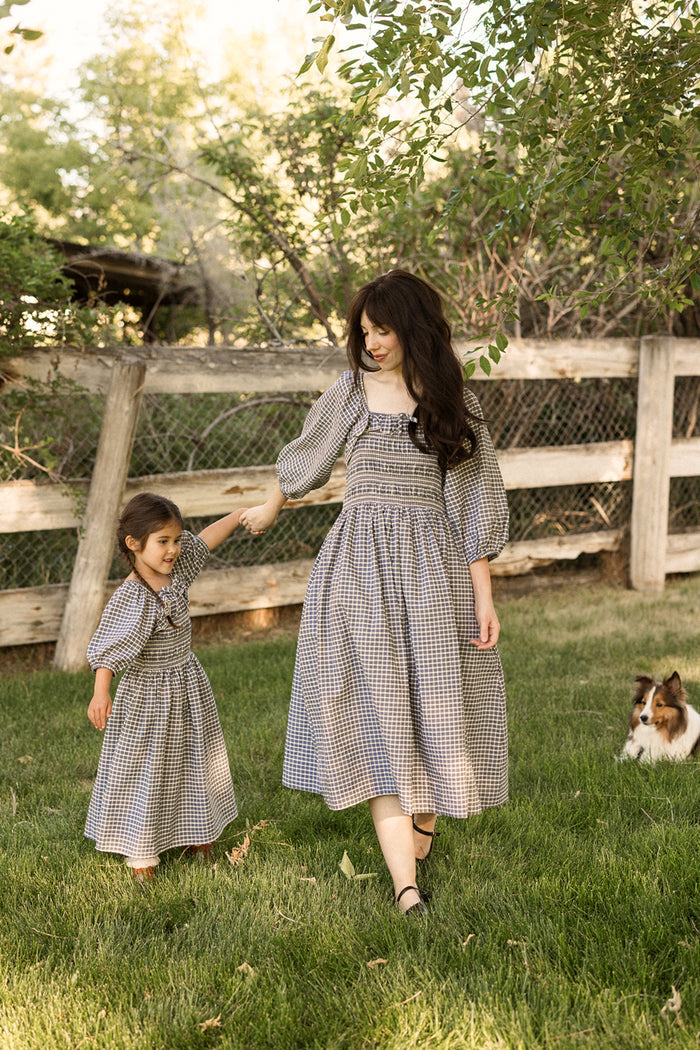 A woman and a young girl, both wearing the Ivy City Co Mini Mae 3/4 Sleeve Dress in Prussian Blue Plaid with a smocked bodice, hold hands and walk on the grass near a wooden fence while a small dog sits nearby beneath the shade of leafy trees.