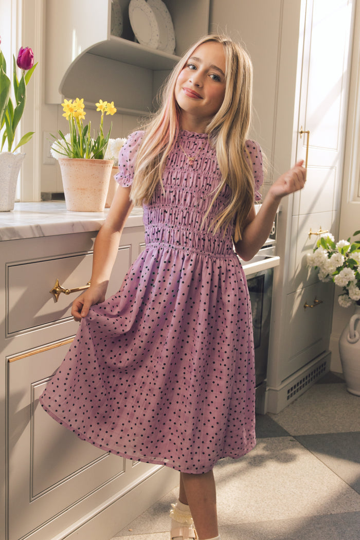 A young girl with long blonde hair wears the Ivy City Co Mini Marcell Dress in Purple Polka Dot, smiling in a sunny kitchen with potted flowers on the counter and background, holding the side of her spring dress.