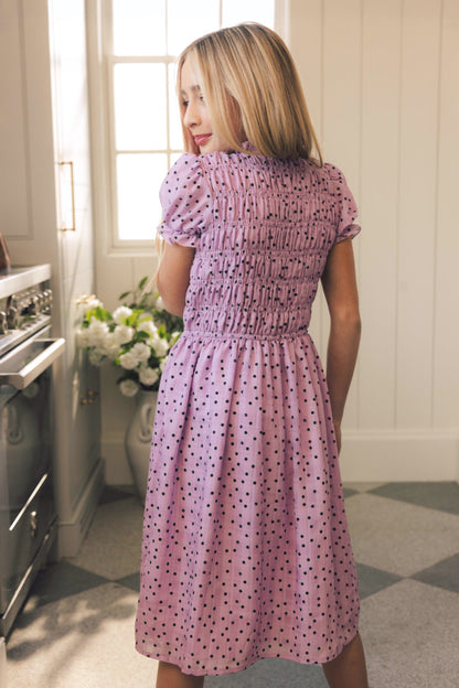 A young woman with long blonde hair stands indoors, sunlight streaming in as she wears the Ivy City Co Mini Marcell Dress in Purple Polka Dot—a ruched, light purple spring dress with a playful polka dot pattern.