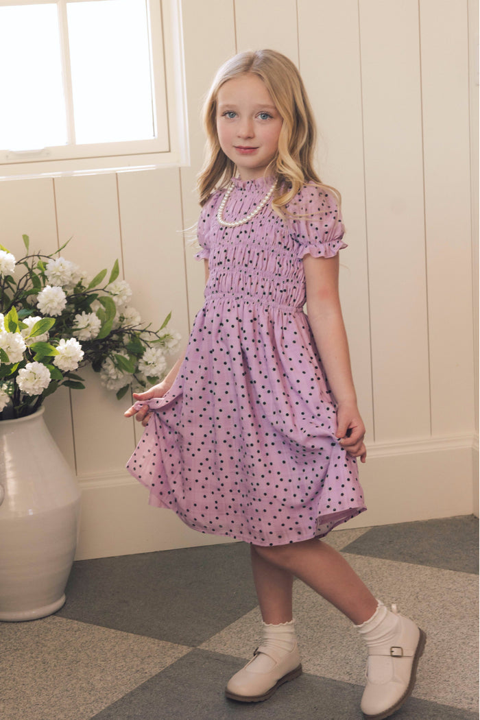 A young girl with long blonde hair wears the Ivy City Co Mini Marcell Dress in Purple Polka Dot, paired with white shoes. She stands indoors by a window and a vase of white flowers, gently holding her dress and smiling at the camera.