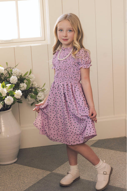 A young girl with long blonde hair wears the Ivy City Co Mini Marcell Dress in Purple Polka Dot, paired with white shoes. She stands indoors by a window and a vase of white flowers, gently holding her dress and smiling at the camera.