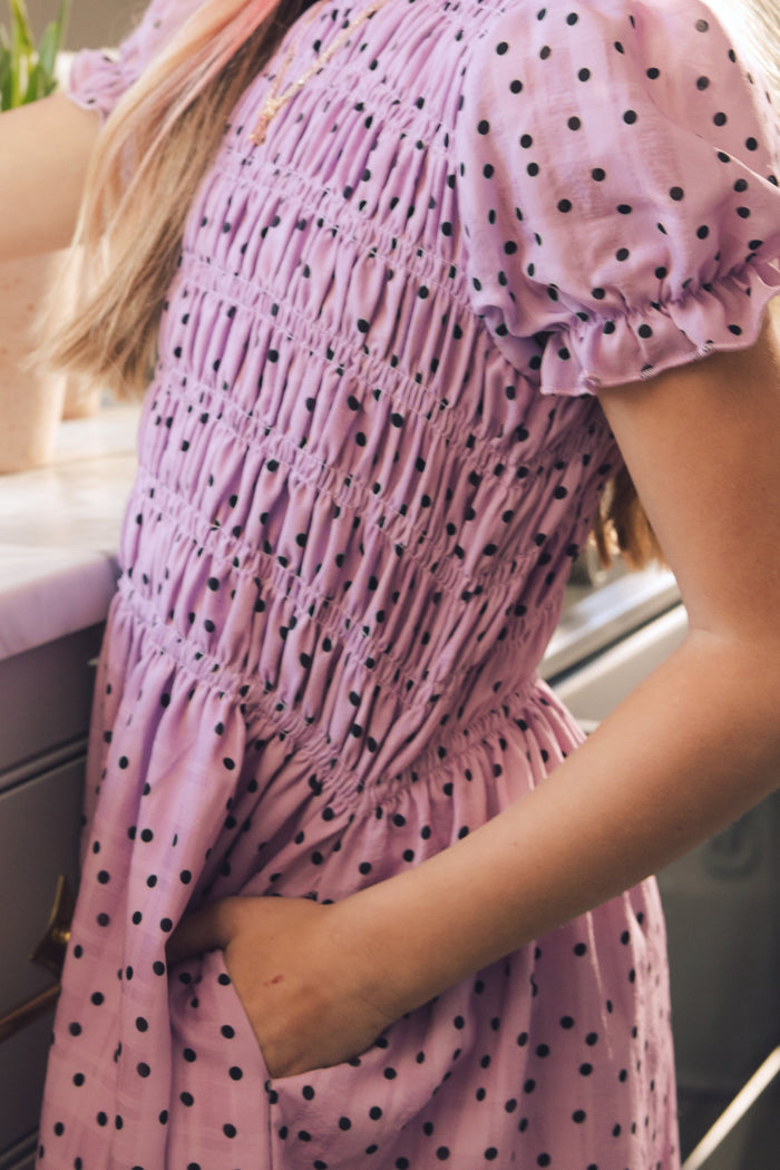 A girl showcases the Ivy City Co Mini Marcell Dress in Purple Polka Dot, with her hand in her pocket. Only her torso and arms are visible, highlighting the playful charm of this classic spring style.