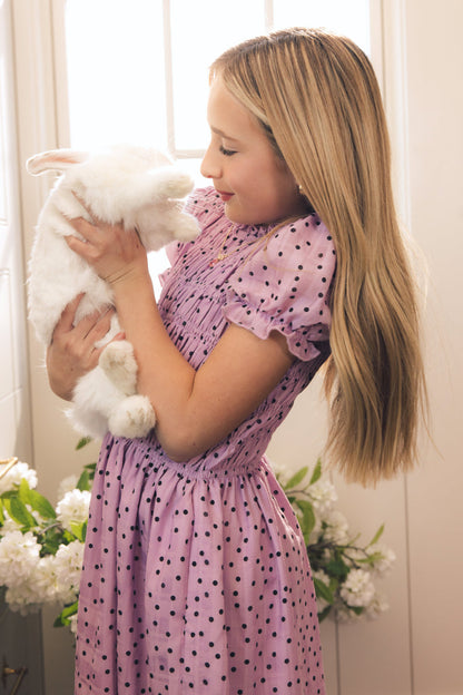 A young girl in an Ivy City Co Mini Marcell Dress in Purple Polka Dot smiles while holding a fluffy white rabbit by a sunlit window, with white flowers in the background.