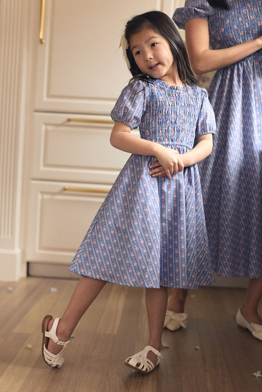 A young girl wears the Ivy City Co Mini Mariah Dress in Linear Print with white sandals, standing on a wooden floor and posing. An adult in a matching dress stands partially visible beside her.