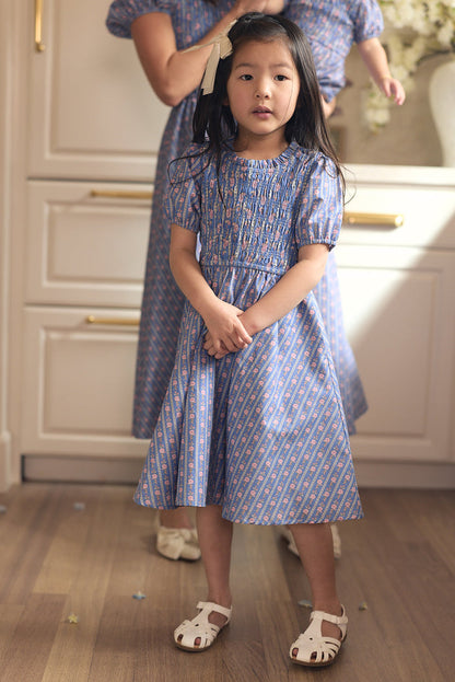 A young girl in the Ivy City Co Mini Mariah Dress in Linear Print, with long dark hair and white sandals, stands indoors on a wooden floor with an adult in a matching dress behind her, framed by light cabinets and soft lighting.
