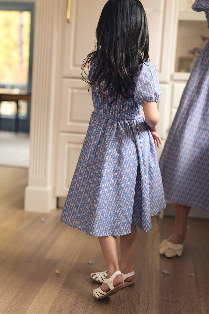 A young girl with long dark hair, wearing the Ivy City Co Mini Mariah Dress in Linear Print and white sandals, stands indoors facing a mirror. An adult in a matching dress appears reflected beside her.