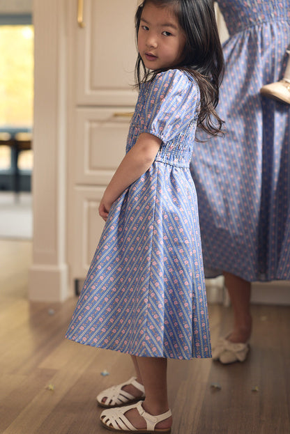 A young girl with long dark hair wears the Ivy City Co Mini Mariah Dress in Linear Print and white sandals, standing indoors on a wooden floor. An adult in a matching dress and white shoes stands partly visible in the background.