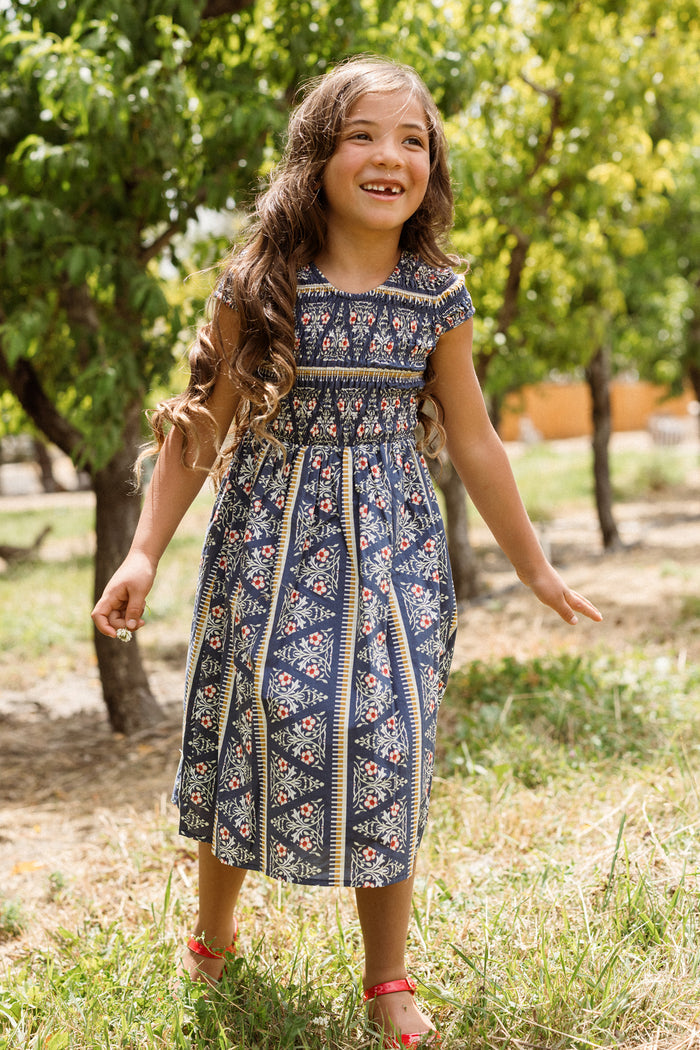 Wearing the Ivy City Co Mini Marlow Cap Sleeve Dress in Navy Fleur, a young girl smiles and plays outside among green trees on a sunny day, her blue patterned dress and red sandals brightening the scene.
