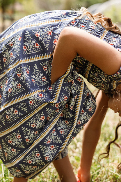 A person wearing the Ivy City Co Mini Marlow Cap Sleeve Dress in Navy Fleur bends down outdoors, with their face partially out of frame and curly hair visible. Grass and greenery are in the background.