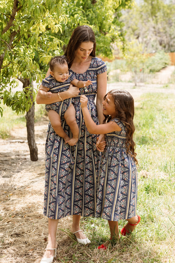 A woman holds a baby and stands next to a smiling young girl. All three wear matching Ivy City Co Mini Marlow Cap Sleeve Dresses in Navy Fleur, featuring delicate patterns that are perfect for sunny days among green trees. The woman’s dress showcases a smocked bodice, adding an elegant touch to their coordinated look.