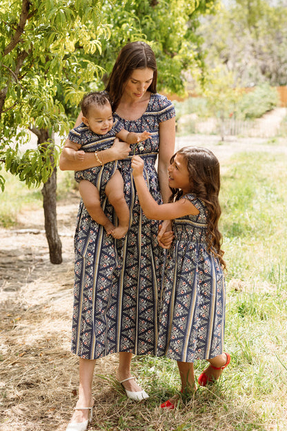 A woman holds a baby and stands next to a smiling young girl. All three wear matching Ivy City Co Mini Marlow Cap Sleeve Dresses in Navy Fleur, featuring delicate patterns that are perfect for sunny days among green trees. The woman’s dress showcases a smocked bodice, adding an elegant touch to their coordinated look.