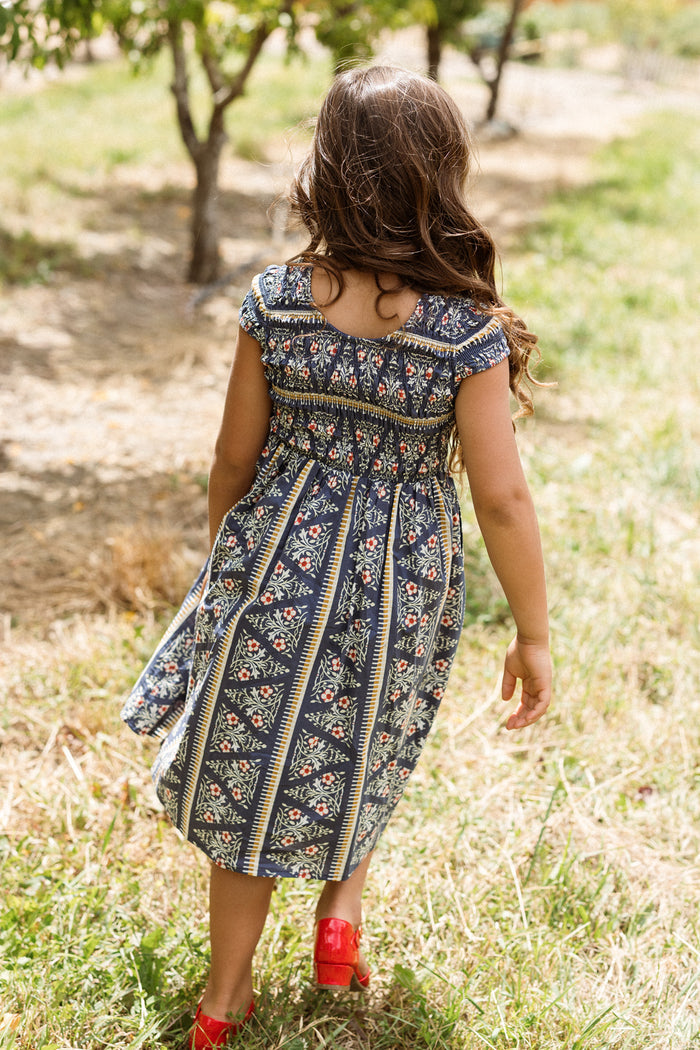 A young girl with long brown hair, wearing the Ivy City Co Mini Marlow Cap Sleeve Dress in Navy Fleur and red shoes, walks away on a grassy path in an outdoor setting with trees.