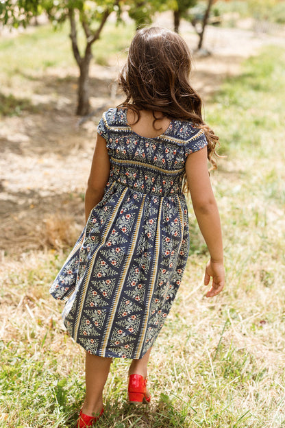 A young girl with long brown hair, wearing the Ivy City Co Mini Marlow Cap Sleeve Dress in Navy Fleur and red shoes, walks away on a grassy path in an outdoor setting with trees.