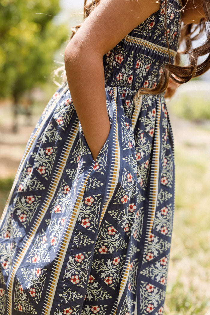A child wearing the Ivy City Co Mini Marlow Cap Sleeve Dress in Navy Fleur stands outdoors with hands in pockets, surrounded by green trees and sunlight.