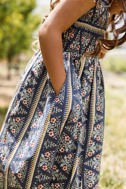 A child wearing the Ivy City Co Mini Marlow Cap Sleeve Dress in Navy Fleur stands outdoors with hands in pockets, surrounded by green trees and sunlight.