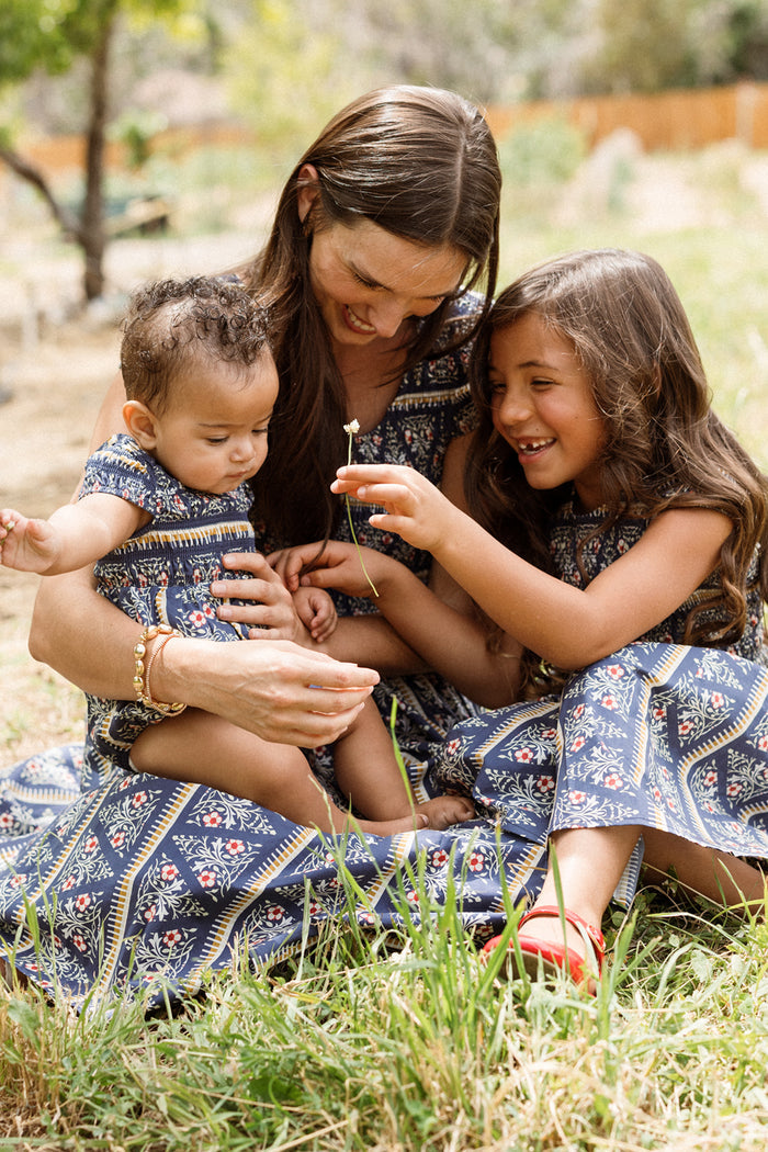 A woman sits on the grass with two young children, all wearing matching Ivy City Co Mini Marlow Cap Sleeve Dresses in the Navy Fleur print. The older child smiles and shows a small flower to the baby while the woman gently supports them.