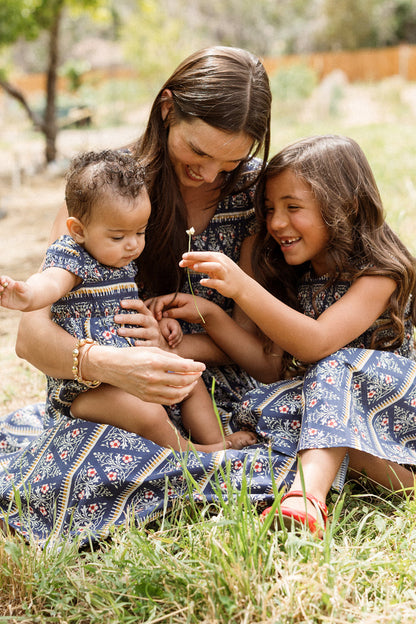 A woman sits on the grass with two young children, all wearing matching Ivy City Co Mini Marlow Cap Sleeve Dresses in the Navy Fleur print. The older child smiles and shows a small flower to the baby while the woman gently supports them.