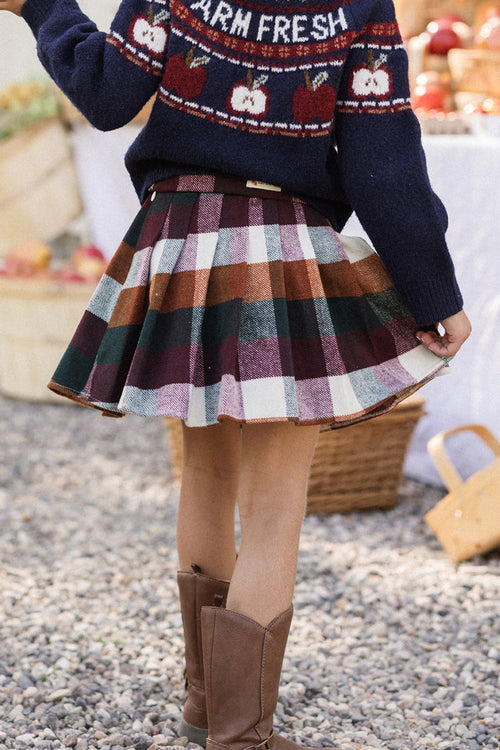 A child wearing a navy FARM FRESH sweater and the Ivy City Co Mini Marren Skirt in Maize Brushed Gingham with pockets stands outdoors on gravel, with brown boots and baskets of apples in the background.