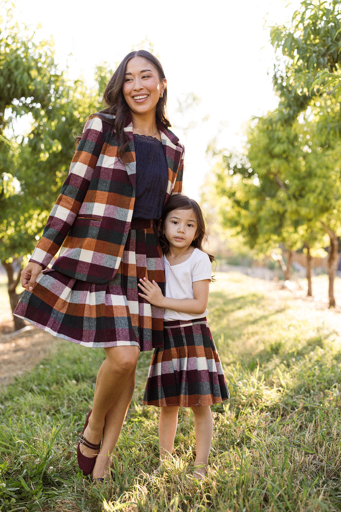 A woman and a young girl stand together on the grass in an orchard, sunlight filtering through the trees. Both wear plaid skirts; the young girl is dressed in Ivy City Co’s Mini Marren Skirt in Maize Brushed Gingham as she hugs the woman’s waist, while the woman pairs her wool-blend skirt with a matching blazer.