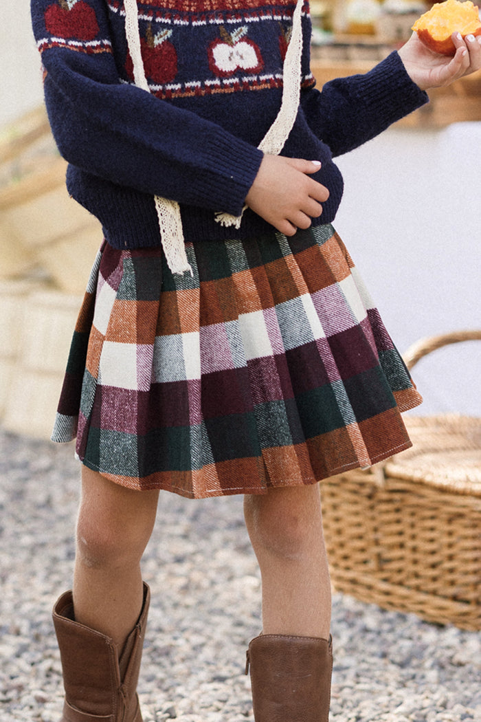 A child wearing a navy sweater with a red and white pattern, the Ivy City Co Mini Marren Skirt in Maize Brushed Gingham, and brown boots stands on gravel while holding an orange fruit. A woven basket can be seen in the background.