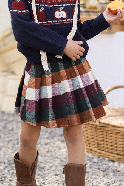 A child wearing a navy sweater with a red and white pattern, the Ivy City Co Mini Marren Skirt in Maize Brushed Gingham, and brown boots stands on gravel while holding an orange fruit. A woven basket can be seen in the background.