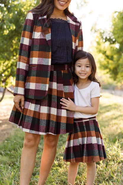 A woman and a young girl stand outdoors on grass with green trees in the background, both wearing matching plaid skirts. The woman completes her look with a coordinating plaid blazer, while the girl beams in the charming Ivy City Co Mini Marren Skirt in Maize Brushed Gingham. Both enjoy the practicality of a skirt with pockets as they embrace and smile together.