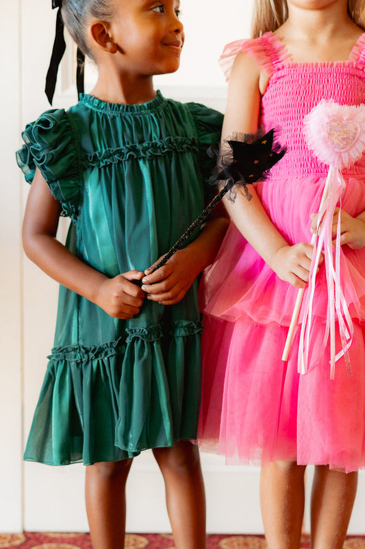 Two young girls stand together—one wears Ivy City Co's Mini Peridot Dress in Green with ruffle sleeves, holding a black star wand; the other is in pink with a ribboned wand. Both look ready for dress-up or a costume party.