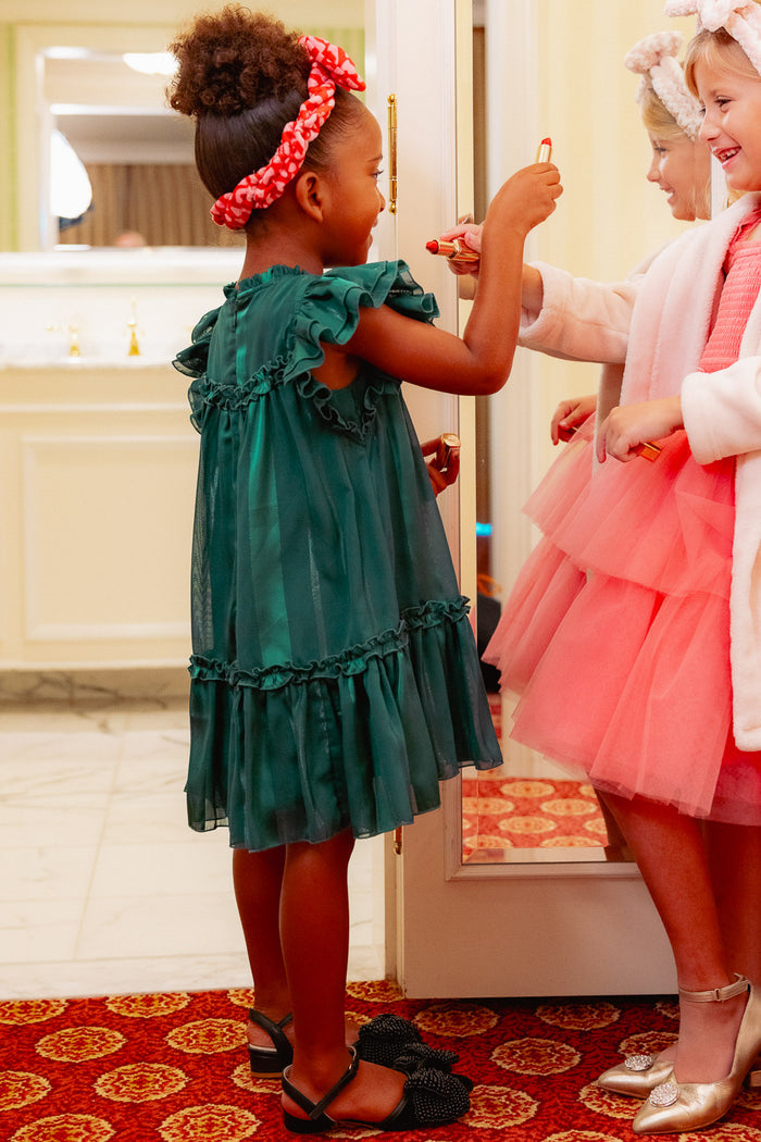 Two young girls smile as they play with lipstick in a bright, elegant room. One wears the Ivy City Co Mini Peridot Dress in Green with ruffle sleeves and a headband; the other sports a pink tulle dress and white fluffy headband.