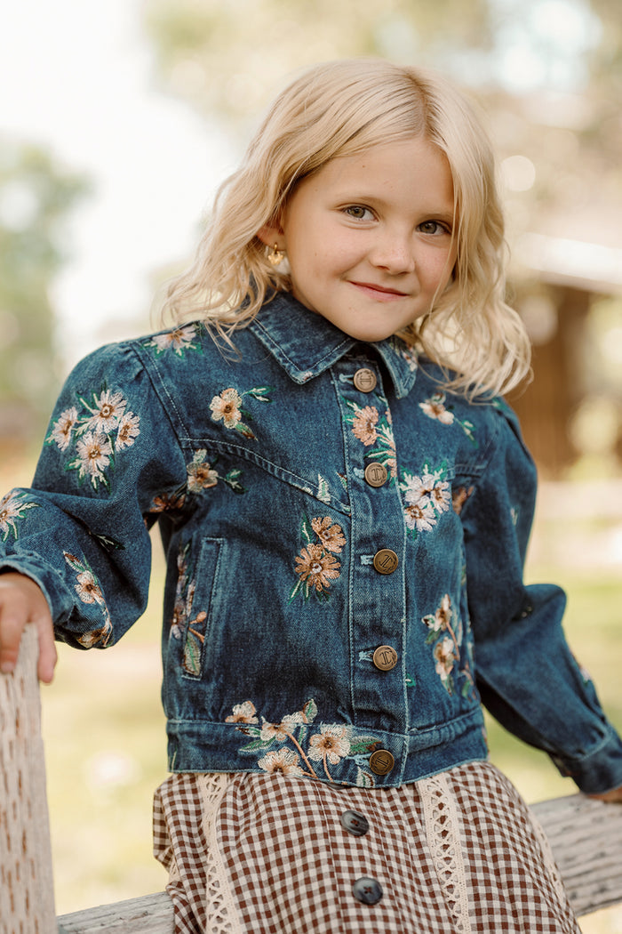 Outdoors, a young blonde girl smiles while wearing Ivy City Co’s Mini Posey Denim Jacket in Lovelorn Ditsy Embroidery layered over a brown gingham dress with lace trim.