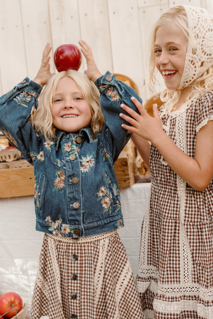 Two young girls with blonde hair wear vintage-inspired outfits—one of them dressed in Ivy City Co’s Mini Posey Denim Jacket in Lovelorn Ditsy Embroidery. Sharing a playful moment, one balances a red apple on her head and beams, while the other laughs and claps her hands. They stand together amidst baskets and wooden crates at what appears to be a market or fair.
