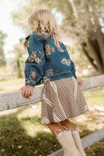 A young child with blonde hair, dressed in the Ivy City Co Mini Posey Denim Jacket in Lovelorn Ditsy Embroidery, a checkered skirt, and white boots, leans on a wooden fence outdoors on a sunny day.