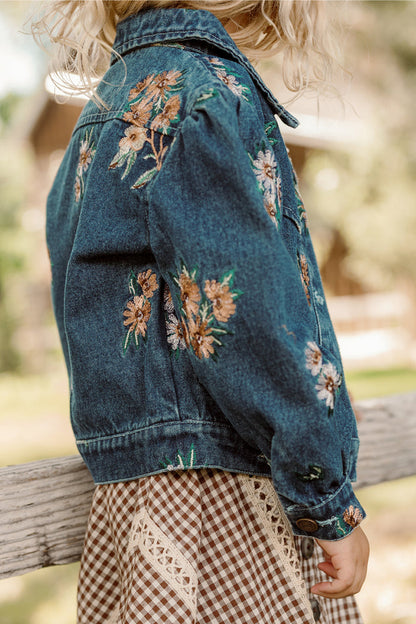 A child with blonde curly hair wears the Ivy City Co Mini Posey Denim Jacket in Lovelorn Ditsy Embroidery and a brown gingham dress with lace trim, standing outdoors near a wooden fence.