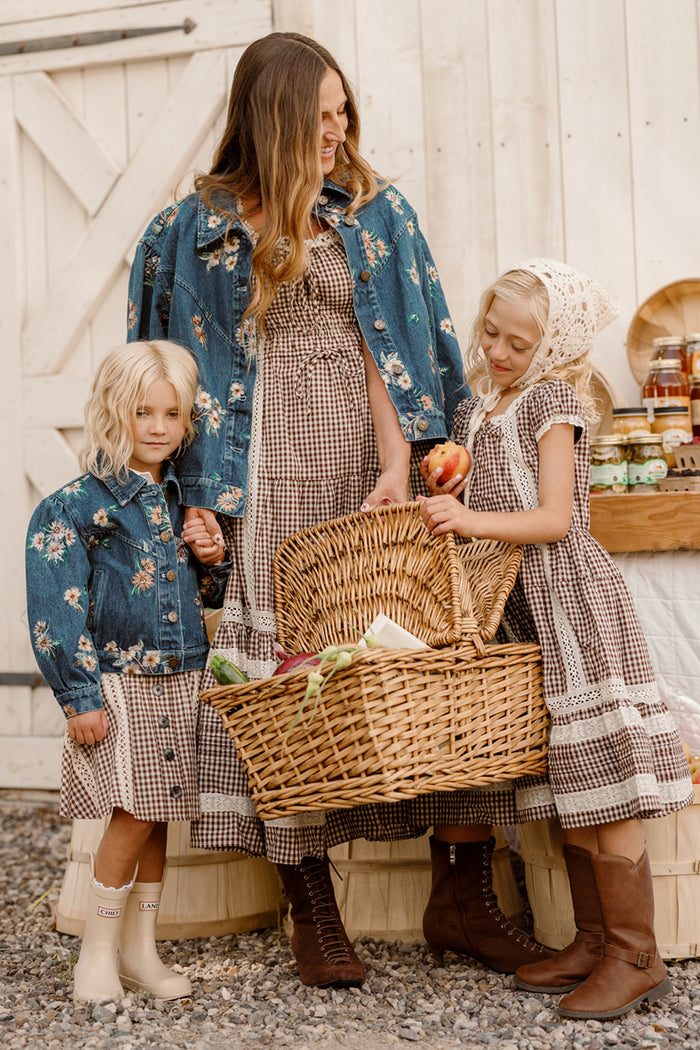 A woman and two young girls in gingham dresses stand by a basket of produce. The woman and one girl are wearing floral denim jackets from Ivy City Co, with the girl showing off the stylish Mini Posey Denim Jacket in Lovelorn Ditsy Embroidery. The other girl, holding an apple and wearing a white headscarf, stands nearby as jars of preserves complete the charming outdoor moment in their girls’ denim jackets.