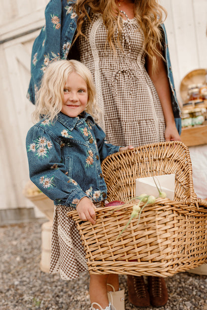 A young girl wearing the Ivy City Co Mini Posey Denim Jacket in Lovelorn Ditsy Embroidery holds a wicker basket filled with vegetables and an envelope, standing beside a woman in a checkered dress in a rustic outdoor setting.