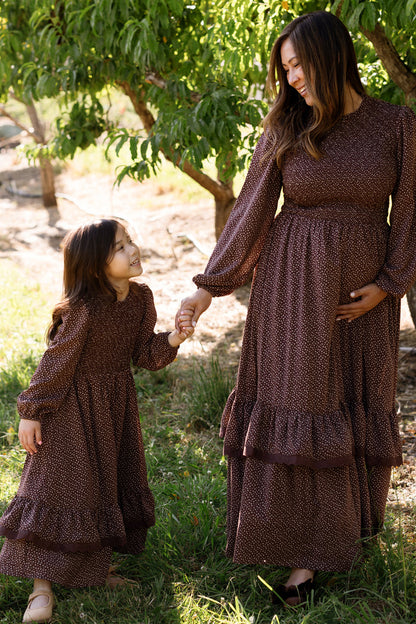 A woman and a young girl, both wearing matching Ivy City Co Mini Presley Maxi Dresses in Mahogany Ditsy with elegant balloon sleeves and a smocked bodice, hold hands and smile at each other while standing outdoors near green trees.