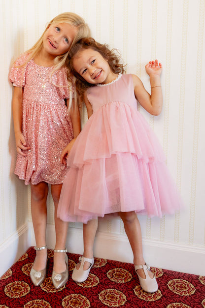 Two young girls smile and pose indoors. One twirls in the Ivy City Co Mini Rainie Dress in Rose—a charming pink tulle special occasion dress—while the other shines in a pink sequined dress and silver shoes. They stand against a light patterned wall on red carpet.