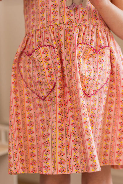 A close-up of the Ivy City Co Mini Rue Play Dress in Handpainted Flora Stripe, a pink sleeveless dress with vertical stripes, small floral prints, and two heart-shaped pockets trimmed with rickrack.