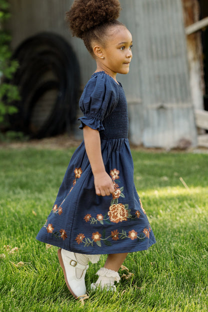 A young girl with curly hair in a bun, wearing the Ivy City Co Mini Teagan Dress in Navy Embroidered Fall Floral and white shoes, walks on green grass beside a corrugated metal building.
