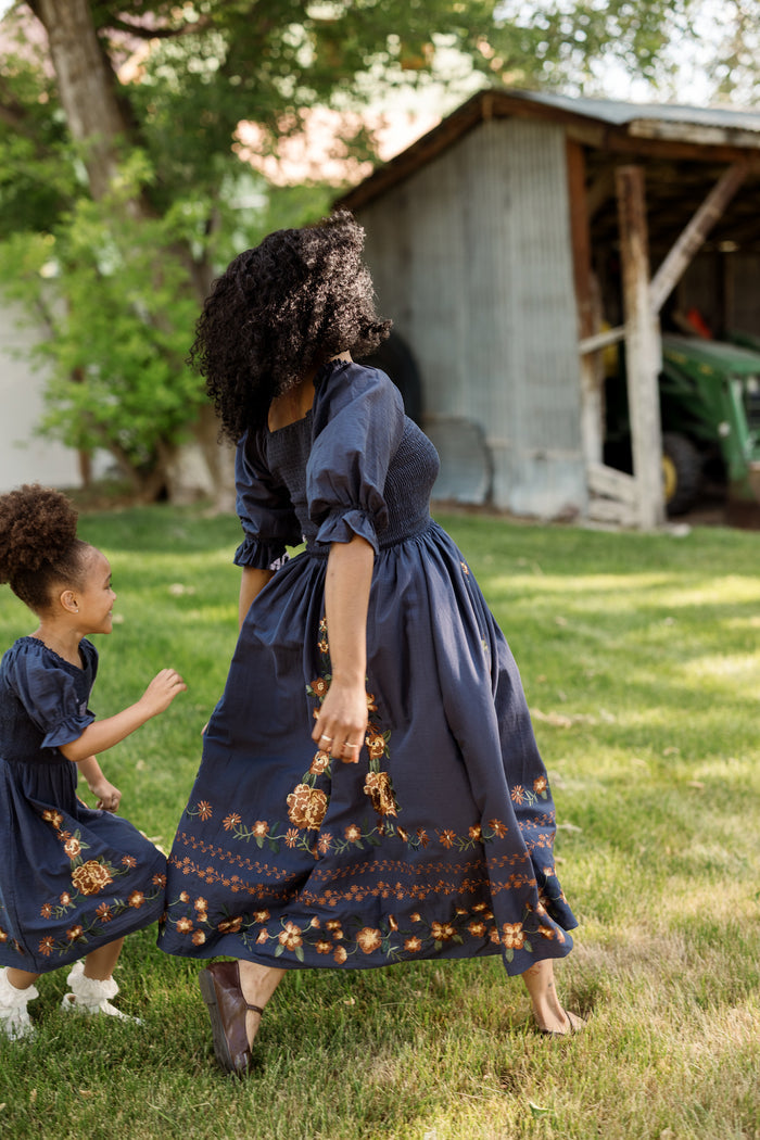 A woman and a young girl, both wearing matching blue cotton dresses with smocked bodices and floral embroidery—such as the Ivy City Co Mini Teagan Dress in Navy Embroidered Fall Floral—hold hands and walk on grass near a rustic shed, surrounded by trees.