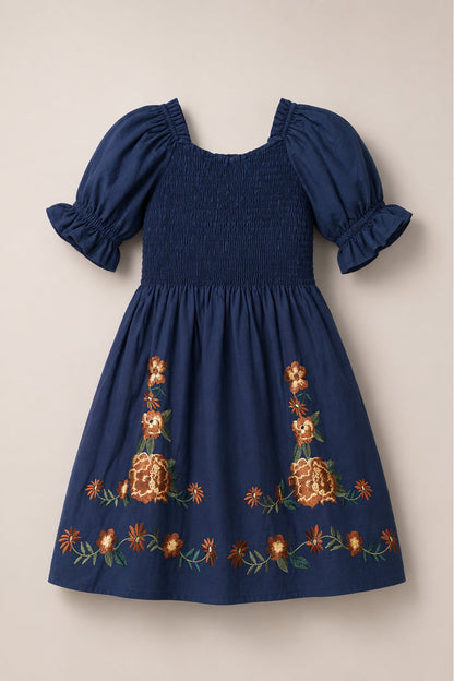 The Mini Teagan Dress in Navy Embroidered Fall Floral by Ivy City Co features a classic navy blue cotton design with puffed sleeves, a smocked bodice, and vibrant orange floral embroidery accenting the hem and center for a playful yet timeless look.