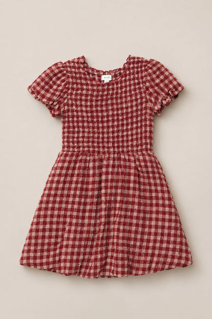 The Mini Tori Dress in Cranberry Popcorn Plaid by Ivy City Co is a short-sleeved, knee-length dress with a red and white checkered pattern, a fitted smocked bodice, and a gathered skirt, displayed laid flat on a neutral background.
