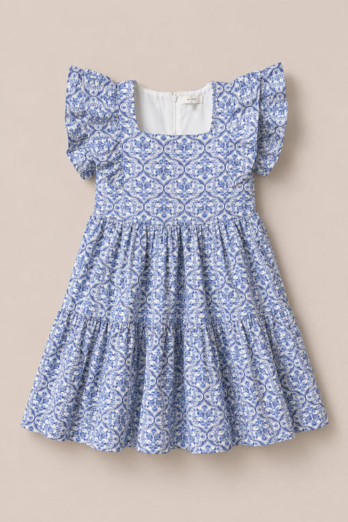 The Mini Trixie Dress in Blue Moroccan Tile by Ivy City Co is a girls' summer dress featuring a blue and white pattern, square neckline, ruffled sleeves, and a tiered skirt, inspired by Moroccan tile designs.