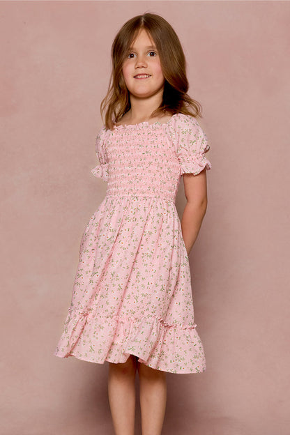 A young girl with light brown hair smiles in front of a pink background, wearing the Ivy City Co Mini Valencia Dress in Pink Ditsy Floral, a charming girls’ midi dress with short ruffled sleeves and her hands behind her back.