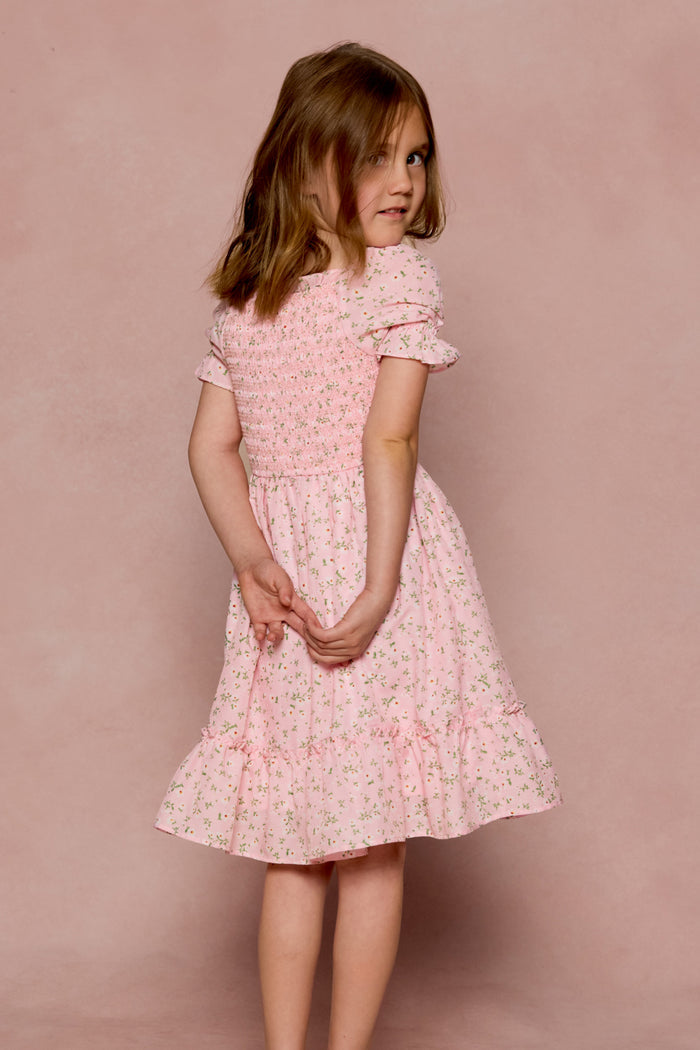 A young girl in the Ivy City Co Mini Valencia Dress in Pink Ditsy Floral stands against a pink background, looking over her shoulder with her hands clasped behind her back.