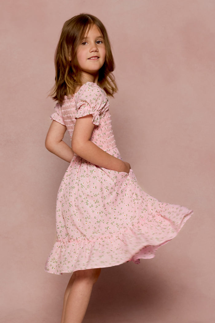 A young girl with light brown hair twirls in the Ivy City Co Mini Valencia Dress in Pink Ditsy Floral, featuring short sleeves and a midi length, against a soft pink background.