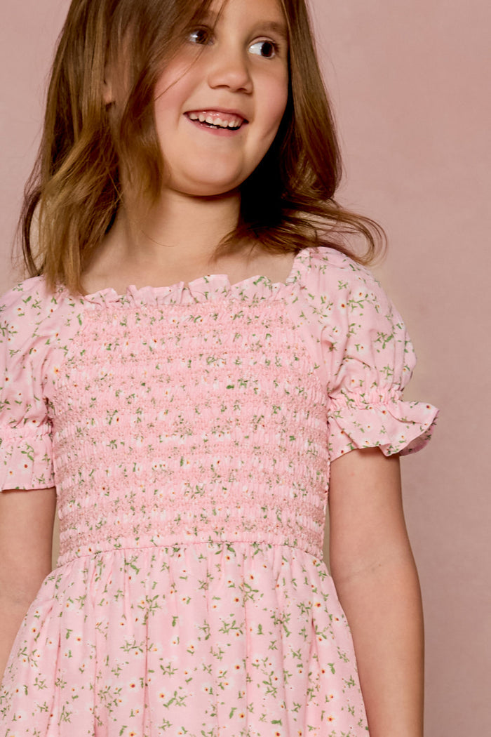 A young girl smiles in the Ivy City Co Mini Valencia Dress in Pink Ditsy Floral, featuring a smocked bodice and short sleeves, as she stands before a matching pink background.